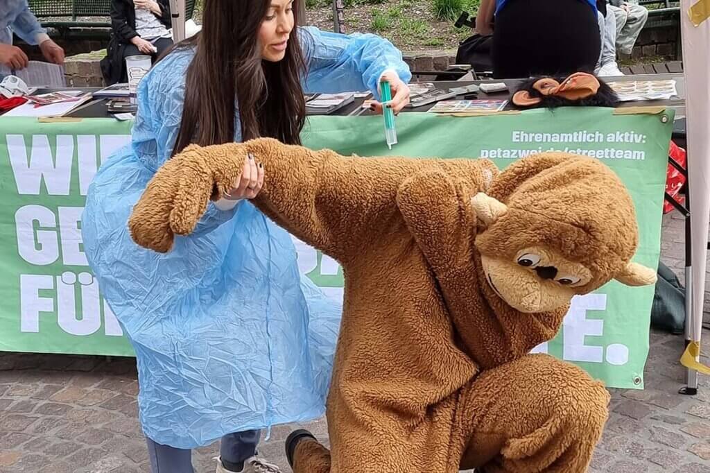 Demonstrierende gegen Tierversuche vor einem Stand. Eine Person im Affenkostuem wird festgehalten von einer Person im Arztkittel und Spritze in der Hand.
