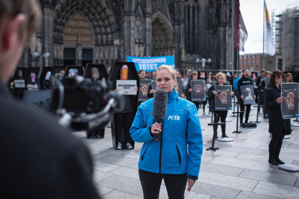 PETA Demo gegen Speziesismus vorm Koelner Dom. Frau in blauer Jacke und Mikrofon in der Hand spricht in eine Kamera.