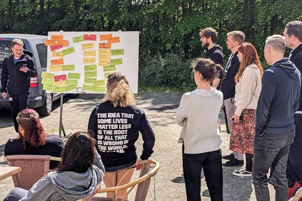 Mehrere Personen stehen auf freiem Gelaende vor einer Pinnwand mit Notizen.