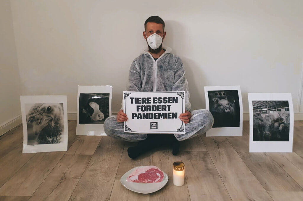 Ein Mensch sitzt auf dem Boden mit Schutzkleidung und Atemmaske und hält ein Schild in der Hand.