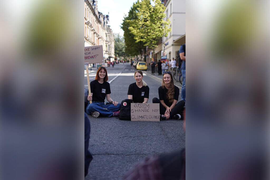 Drei Menschen sitzen auf der Straße auf dem Boden und halten Plakate in der Hand.