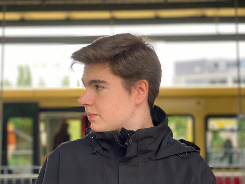 Milan steht an einem bahngleis schaut von sich aus nach rechts und trägt dabei eine Kurzhaarfrisur und eine schwarze Windbreaker-Jacke.